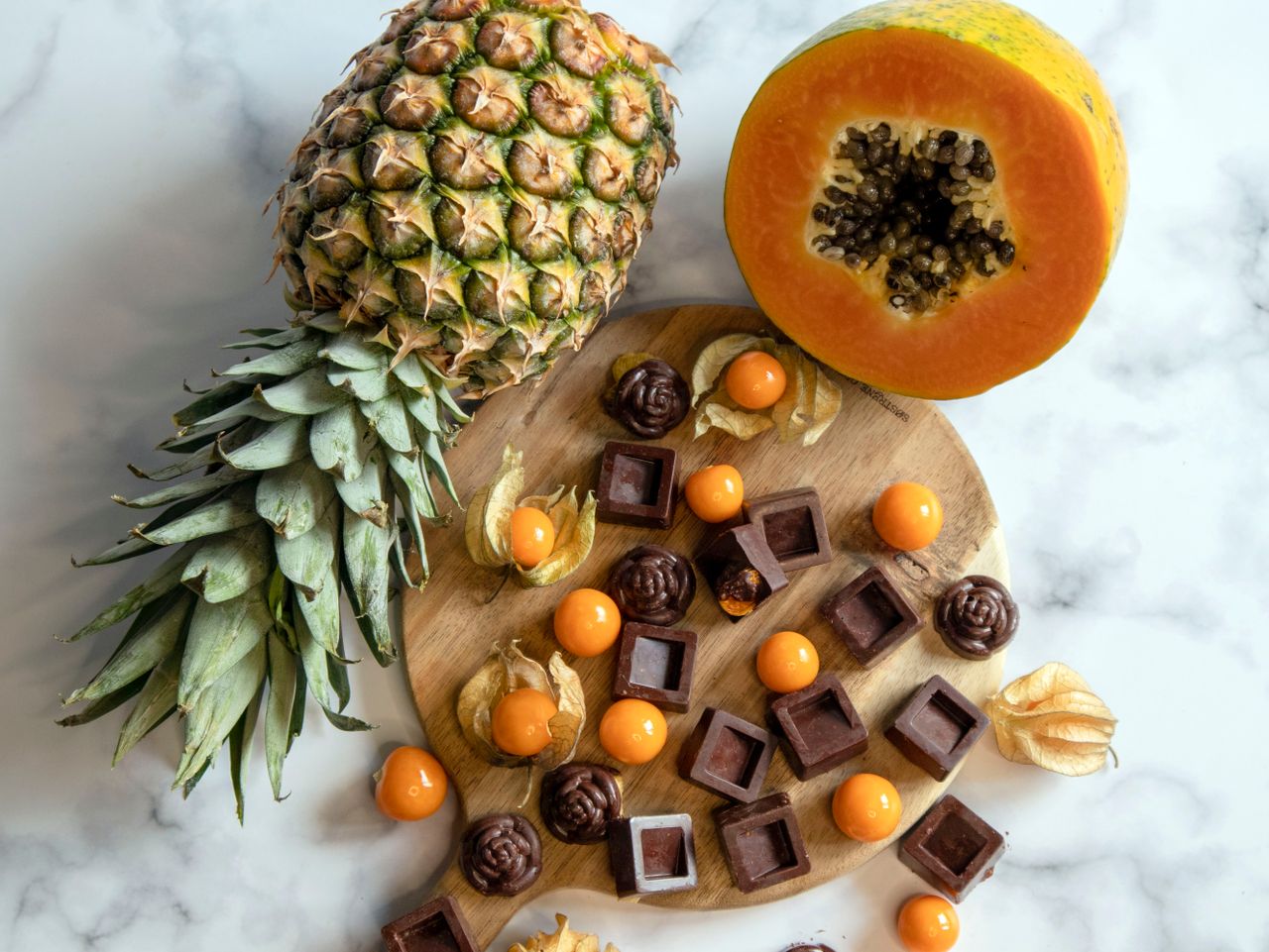 A vibrant arrangement of pineapple, papaya, and assorted chocolates on a wooden board and marble surface.